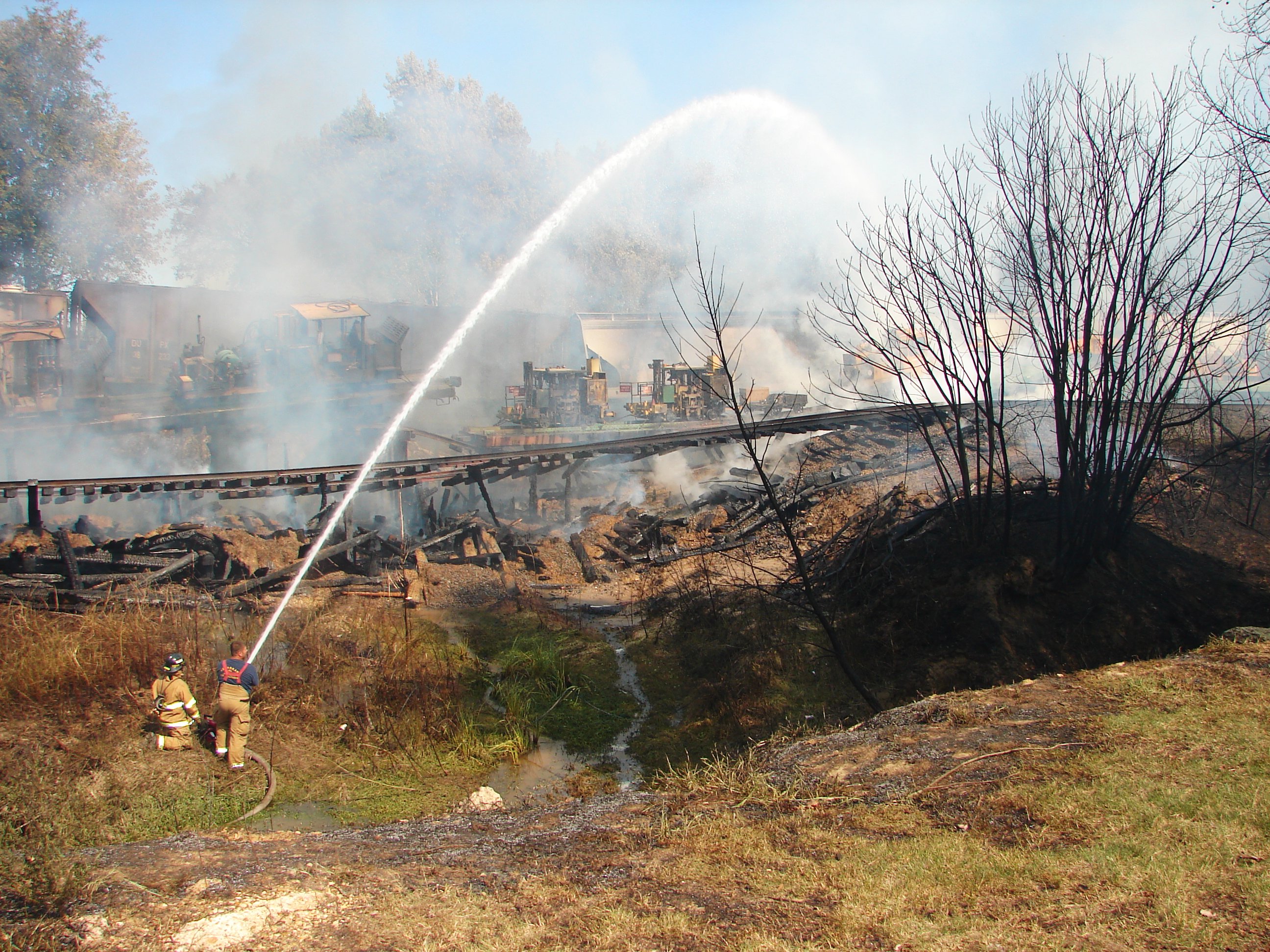 Two fire fighters shoot a stream of water onto the fire, over the train tracks.